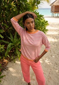 Mujer con camisa de rayas rosas y pantalones coral de pie en un sendero de arena junto a plantas tropicales y una choza de playa, mirando de lado con la mano en la cabeza.