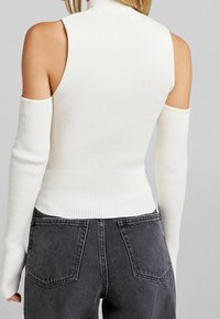Top bianco a coste senza maniche con scollo alto e maniche lunghe. Abbinato a jeans grigio scuro a vita alta con tasche posteriori.