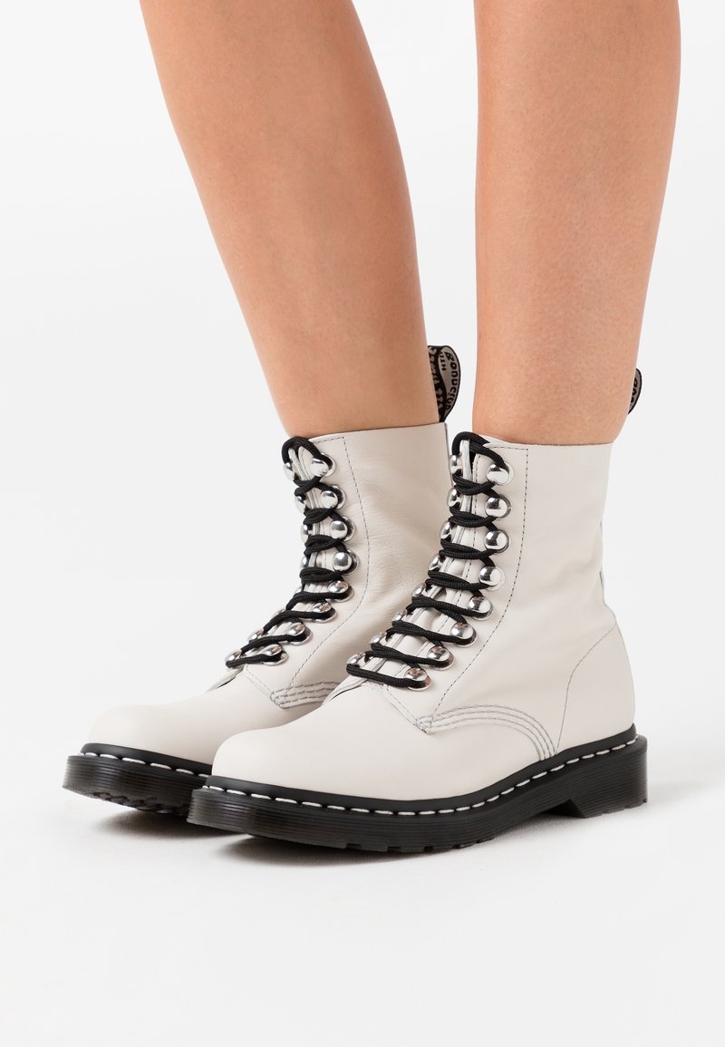 Bottes blanches en cuir à lacets avec semelles noires et lacets noirs portées sur des jambes nues sur fond blanc.