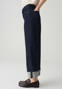 Jeans in denim blu scuro con una vestibilità rilassata, orlo grigio risvoltato e tasche frontali. Abbinati a scarpe di pelle marrone.