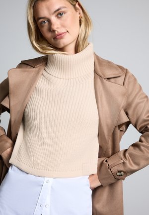 Un pull col roulé beige à côtes associé à un trench-coat marron clair, avec un col cranté et des détails de poignets boutonnés. Le fond est gris.
