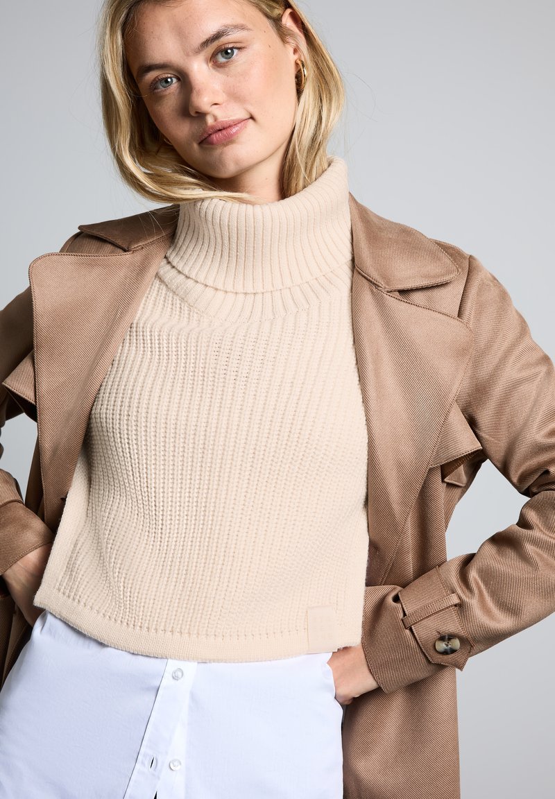 Beżowy sweter z prążkowanym golfem w połączeniu z jasnobrązowym płaszczem trench, z wyciętym kołnierzem i guzikami na mankietach. Tło jest szare.