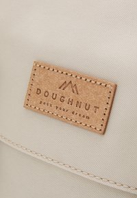 Beige fabric backpack featuring a textured brown leather patch with "DOUGHNUT" embossed, and stitching details along the edges.