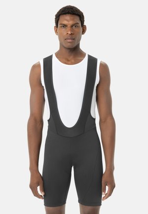 Mannelijk model draagt zwarte wielerbroek met bretels en een witte mouwloze basislaag, staand tegen een effen lichte achtergrond.