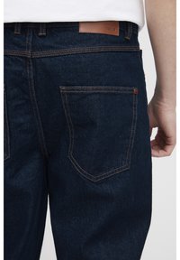 Jeans de mezclilla azul oscuro con costuras naranjas, con un bolsillo trasero y una etiqueta de cuero marrón en la cintura. Superficie texturizada visible.