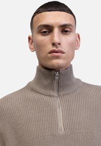 Neule, puolivetoketjullinen pullover taupe-värissä, jossa on ribbitekstuuri ja korkea kaulus. Vetoketjussa on metallinen viimeistely. Lähempänä näkyvä kuva vaatteesta.