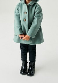 Manteau en polaire vert menthe avec capuche, trois boutons beige et poches latérales, associé à un jean skinny noir et des bottines noires brillantes.