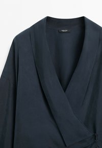 Tmavě námořnický zavinovací oděv s šálovým límcem a bočními knoflíky, zobrazený na bílém pozadí.