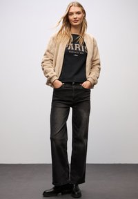 Beige, fuzzy bomberjakke over en sort sweatshirt med "PARIS" tekst, parret med sorte straight-leg jeans og sorte ankelstøvler.