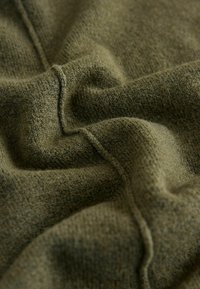 OTSZYW KORALOWITTY CZARNO-OLIWKOWEJ Z DZIANINY CHARAKTERYZUJĄCEJ SIĘ MIĘKKĄ, STRUKTURALNĄ POWIERZCHNIĄ Z WIDOCZNYMI SZWAMI. ZGUBY TKANINY TWORZĄ RÓŻNORODNE WZORY ŚWIATŁA I CIENIA.