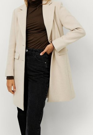 Manteau beige mi-genoux porté sur un col roulé marron et un jean noir taille haute, avec une main dans la poche et une posture détendue.