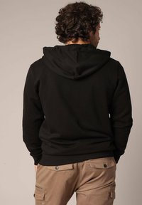Zwarte hoodie met een zachte textuur en een verstelbare trekkoordcapuchon. Gecombineerd met tan kleurige cargobroek met zakken en knoopaccenten.