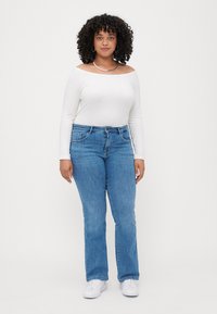 Haut blanc à manches longues décontracté et décolleté porté avec un jean évasé en denim bleu. Le modèle porte des baskets blanches ; ses cheveux bouclés et un collier sont visibles.