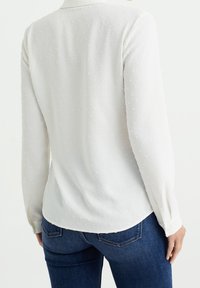 Weiße Bluse mit strukturiertem Muster, langen Ärmeln und geknöpften Manschetten, kombiniert mit dunkelblauen Jeans. Die Rückansicht zeigt einen abgerundeten Saum.