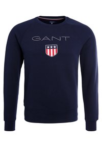 Felpa blu navy con maniche lunghe a raglan. Presenta il logo "GANT" e uno scudo con strisce rosse, bianche e blu. Realizzata in tessuto morbido e testurizzato.