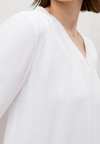 Blusa bianca a maniche corte con scollatura a V e un sottile motivo testurizzato. Il tessuto appare leggero e liscio.