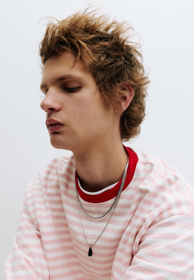 Jeune personne aux cheveux châtain clair en bataille, portant une chemise rayée rose et blanche avec un col rouge, et deux colliers en argent avec un pendentif.
