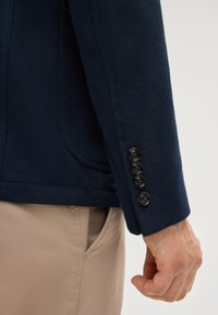 Marineblaue strukturierte Jacke mit einer Nahaufnahme eines Ärmels, der vier schwarze Knöpfe und ein dezentes Taschen-Detail zeigt. Beigefarbene Hose sichtbar.