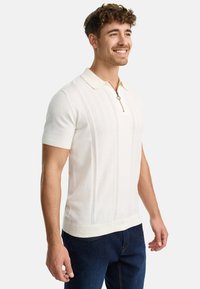 Polo blanco de manga corta con cuello de cremallera, que presenta patrones de punto texturizados y un cuello claro en contraste. Combinado con jeans azul oscuro.