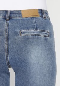 Ljusblå denimjeans med en strukturerad yta, med en rektangulär brun etikett i midjan och en liten bakficka.