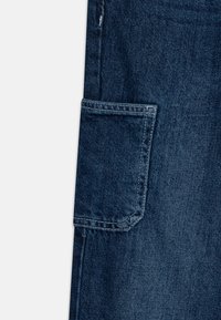 Mørkeblå denimstoff med en stor sidelomme med forsterket søm og en falmet tekstur. En rettskåret ben design er synlig.