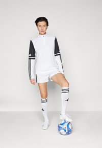 Hvid atletisk trøje med sorte ærmer, der har tre striber og en halvlange lynlås krave, parret med hvide shorts og knæhøje sokker. Blå fodbold.