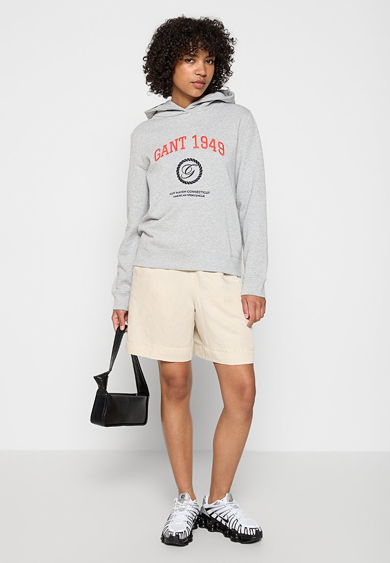 Grauer Kapuzensweatshirt mit "GANT 1949" in Rot, beigen Shorts, schwarzer Handtasche und weißen Sneakers mit schwarzen Akzenten. Leger Outfit.