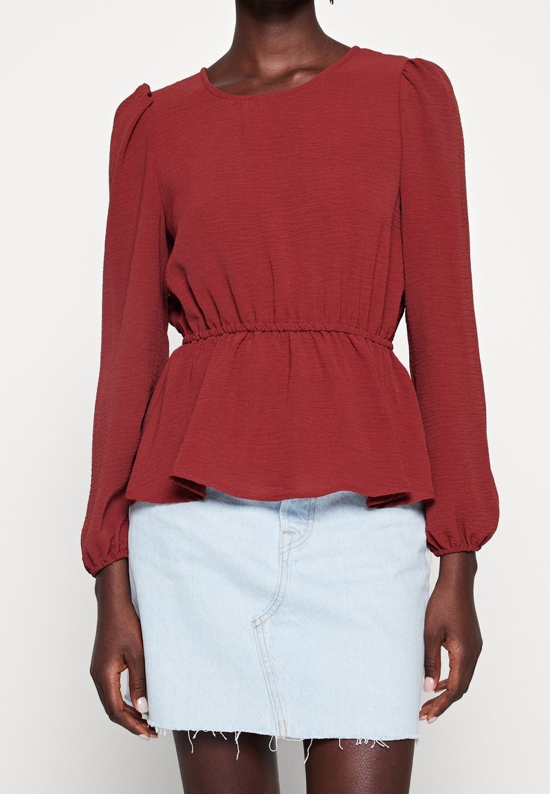 Femme portant un chemisier rouge rouille à manches longues avec une taille élastique et une mini-jupe en jean bleu clair effilochée sur fond blanc.