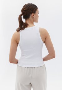 Weißes, geripptes Tanktop mit figurbetonter Passform und breiten Schulterträgern, von hinten vor einem hellen, einfarbigen Hintergrund gezeigt.