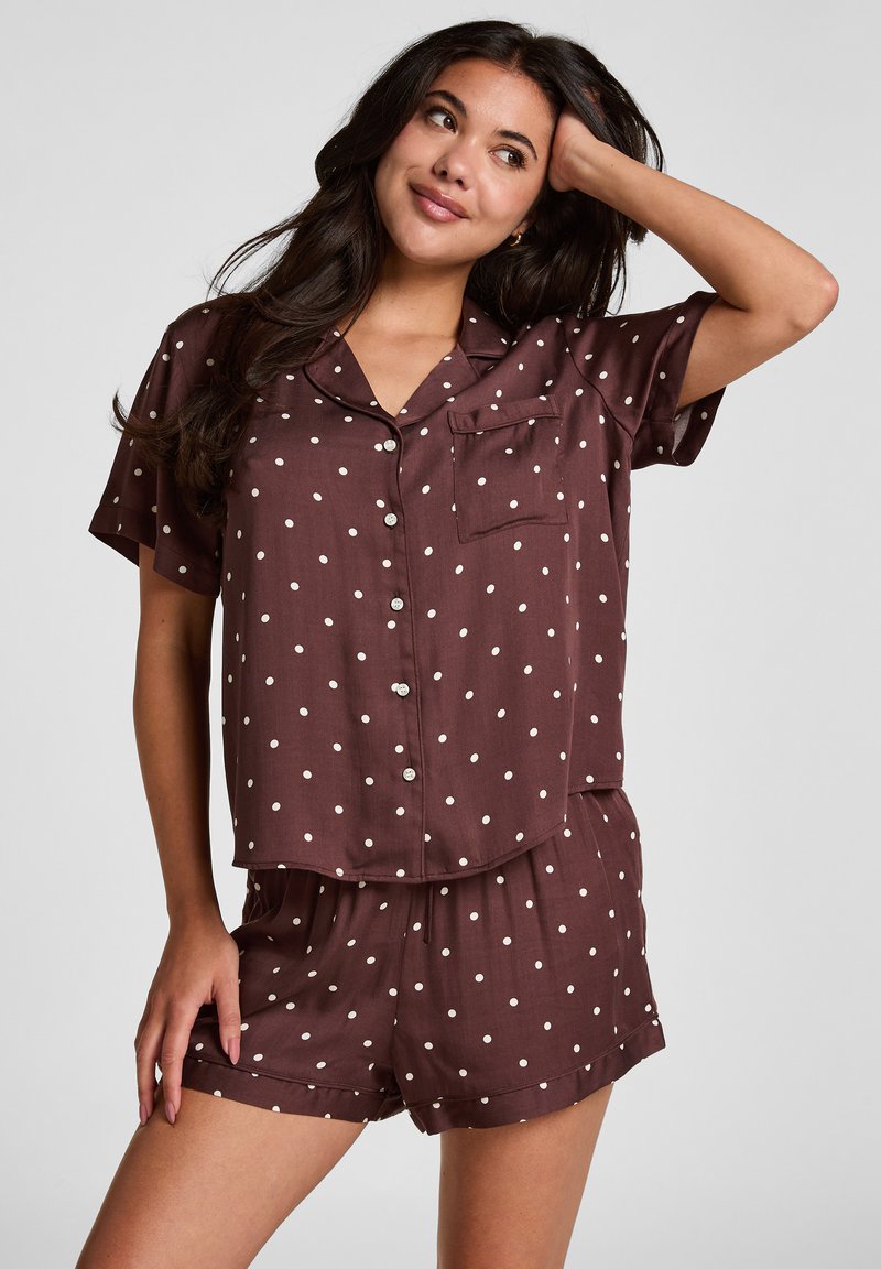 Femme portant une chemise marron à manches courtes avec boutons et un short assorti à pois blancs, posant avec une main sur la tête.