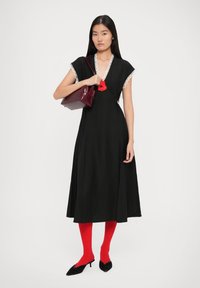 Vestido negro con encaje en el dobladillo, silueta A-line y un acento de flor roja. La modelo sostiene un bolso burdeos, lleva medias rojas y zapatos negros.
