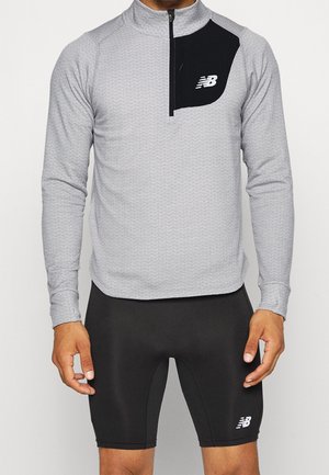 Hombre vistiendo una camiseta gris claro de manga larga con cremallera en el cuarto superior, con panel negro en el pecho y pantalones cortos deportivos ajustados negros con logotipos.