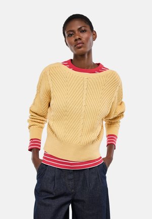 Femme portant un pull jaune à torsades par-dessus une chemise à rayures rouges et blanches et un pantalon bleu foncé, debout les mains dans les poches.