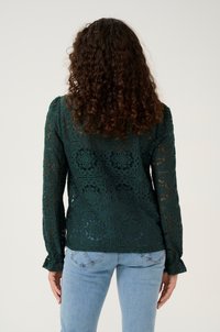Green lace blouse with floral patterns, long sleeves, textured finish, and fitted design, paired with light blue jeans.