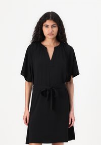 Μαύρο φόρεμα μέχρι το γόνατο με κοντά μανίκια, V-λαιμόκοψη και ζώνη που δένει στη μέση. Λείος υφασμάτινος χαρακτήρας, απλός σχεδιασμός, χωρίς σχέδια ή αξεσουάρ.