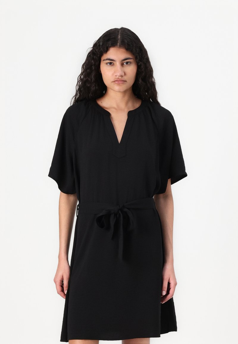 Μαύρο φόρεμα μέχρι το γόνατο με κοντά μανίκια, V-λαιμόκοψη και ζώνη που δένει στη μέση. Λείος υφασμάτινος χαρακτήρας, απλός σχεδιασμός, χωρίς σχέδια ή αξεσουάρ.