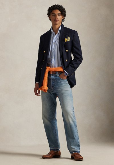 Δίγαζο μπλέιζερ ναυτικού τύπου, ριγέ πουκάμισο, πορτοκαλί πουλόβερ δεμένο στη μέση, ανοιχτό μπλε τζιν, καφέ παπούτσια. Το μοντέλο στέκεται με τα χέρια στις τσέπες.