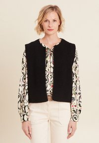 Schwarze gestrickte Weste mit offenem Vorderteil, Strukturstoff, getragen über einer mehrfarbigen gemusterten Bluse. Helle beigen Hosen sind sichtbar.