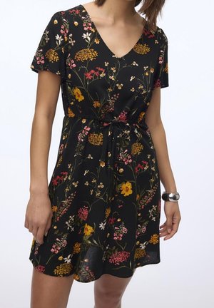 Femme portant une robe noire à manches courtes et col en V, imprimé floral coloré avec une ceinture à la taille, accessoirisée d'un bracelet en argent.