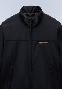 Veste noire zippée avec col montant et poches latérales, arborant un écusson rectangulaire du logo Napapijri Geographic sur la poitrine gauche.