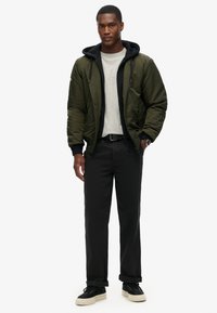 Giacca bomber di colore verde oliva, con cappuccio nero, abbinata a una maglietta grigia chiara e pantaloni neri. Le scarpe sono nere con suola bianca.