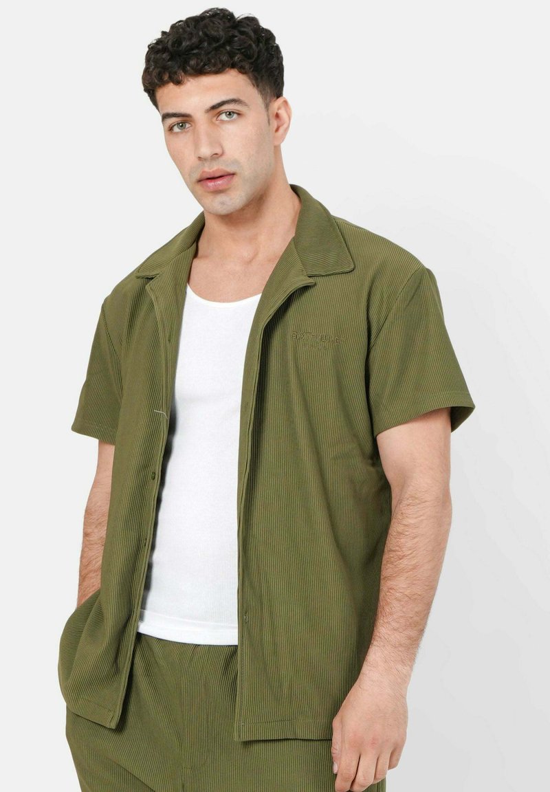 Korte mouwen, olijfgroen gestreepte blouse met een ingeknipte kraag. Draagt over een witte tanktop, heeft een ontspannen pasvorm en voorzakken.