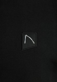 Tessuto nero lavorato a maglia con una piccola etichetta quadrata che presenta un disegno astratto bianco. La texture è liscia e uniforme. Nessun motivo o accento aggiuntivo.