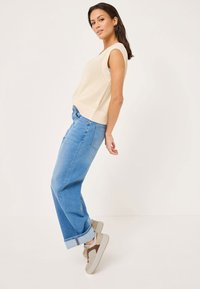 Beige ribbad ärmlös topp med en avslappnad passform, kombinerad med ljusblå vida jeans och tvåtoniga vardagssneakers, allt mot en neutral bakgrund.