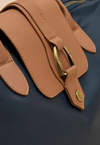 Bruine getextureerde leren flap met een gouden metalen sluiting op een donkere stoffen achtergrond. Heeft afgeronde hoeken en stiksel details.