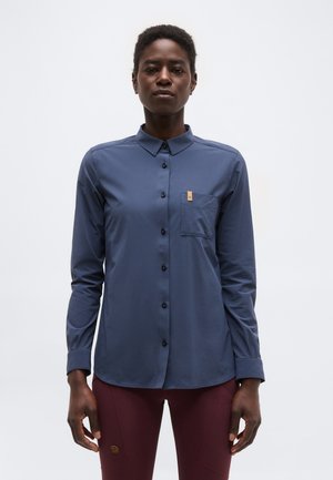 Άτομο ντυμένο με μακρυμάνικο μπλε πουκάμισο με κουμπιά και τσέπη στο στήθος, καθώς και βυσσινί παντελόνι, στέκεται μπροστά σε ένα απλό λευκό φόντο.