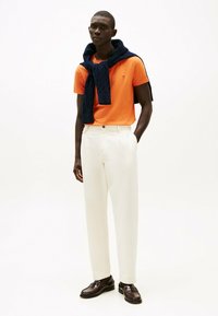 Polo shirt arancione a maniche corte con un maglione in maglia blu navy drappato sulle spalle, abbinato a pantaloni bianchi su misura e scarpe marroni.