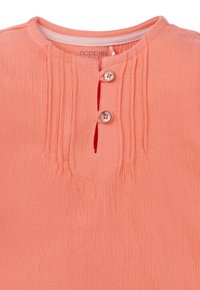 Haut texturé de couleur pêche avec un col rond, deux boutons de fermeture et de subtils plis. Tissu doux avec un design léger.