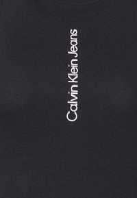Svart bomullsskjorta med vertikal vit text som säger "Calvin Klein Jeans" i en modern sans-serif typsnitt. Enkel, odekorerad design.