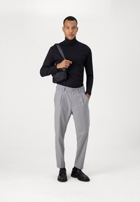 Pull noir à col roulé, pantalon grisé à plissés, et sac bandoulière noir. Le modèle porte des chaussures noires. Fond simple, tons neutres.